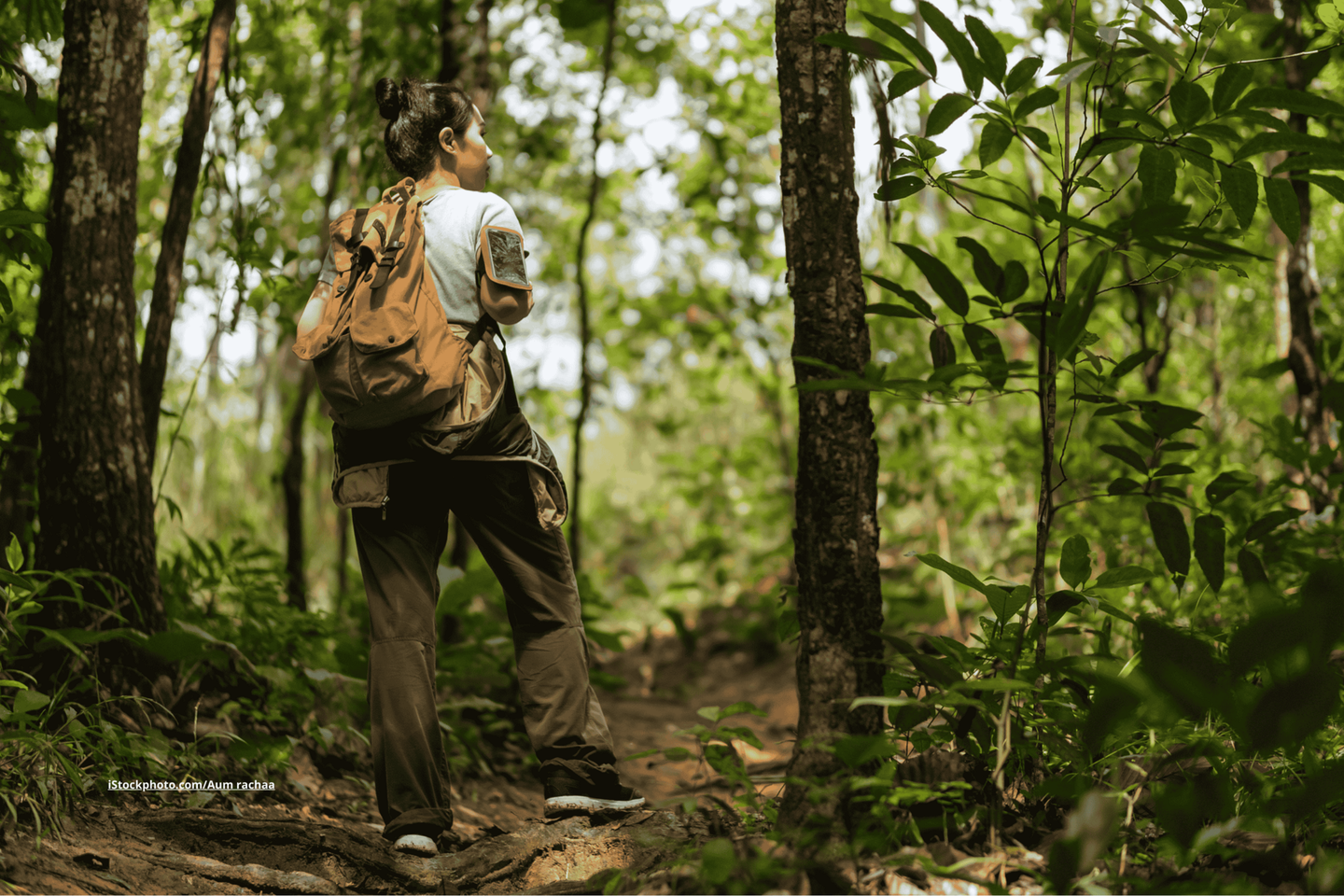 Eine Frau, die durch einen Wald läuft. Sie steht mit dem Rücken zur Kamera und trägt eine braune Hose, ein helles T-Shirt und einen senfgelben Rucksack. 