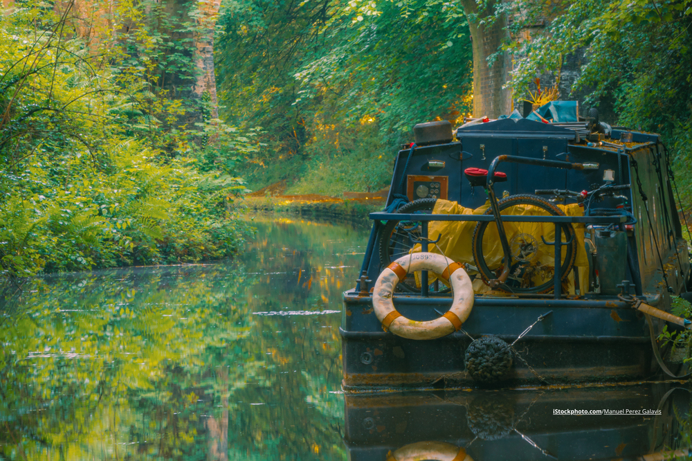 Ein Hausboot, das auf einem Fluss schwimmt. Im Hintergrund sind eine Brück eund Bäume zu sehen. 