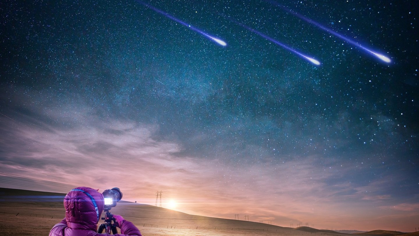 Mensch mit pinker Jacke und Kamera auf Stativ fotografiert Sternschnuppenschauer am Nachthhimmel.