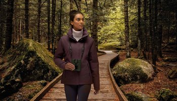 Das Foto zeigt eine Frau mit Fernglas auf einem Spaziergang im Wald. 