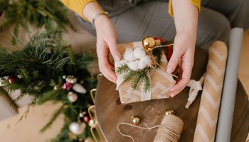 Zwei Hände halten ein weihnachtlich verpacktes Geschenk auf einem Tisch neben einem Tannenbaum.