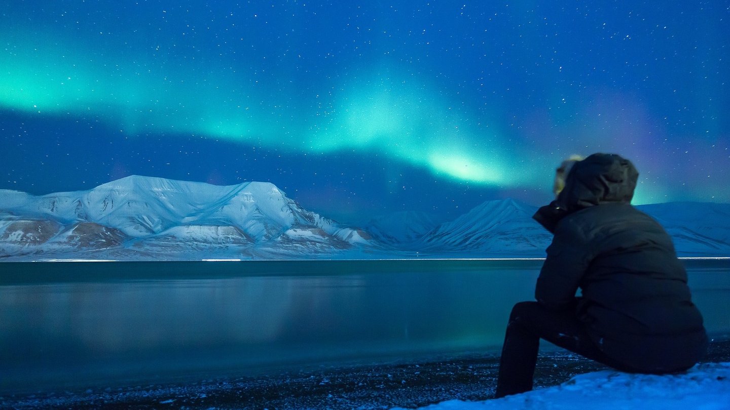 Eine Person sitzt am Ufer und beobachtet die magischen Polarlichter, die über verschneite Berge und einen Sternenhimmel tanzen.