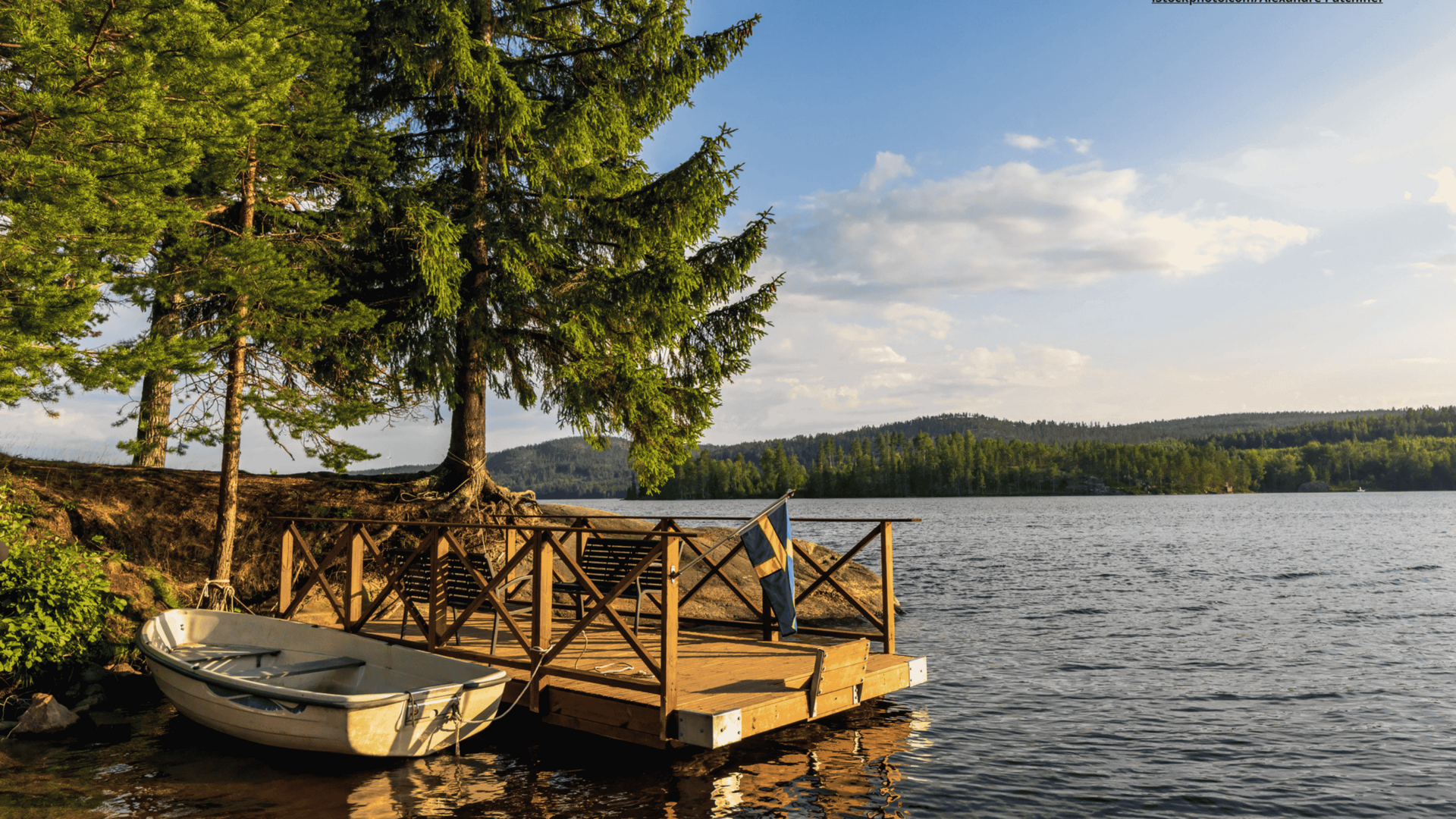 Es ist ein kleines, weißes Boot neben einem Holzsteg im Wasser zu sehen. Der Steg führt an Land, wo mehrere grüne Bäume stehen. Im Vordergrund und Hintergrund des Bildes ist Wasser zu sehen. In der Distanz sieht man eine hügelige Waldlandschaft. Der Himmel ist blau. 