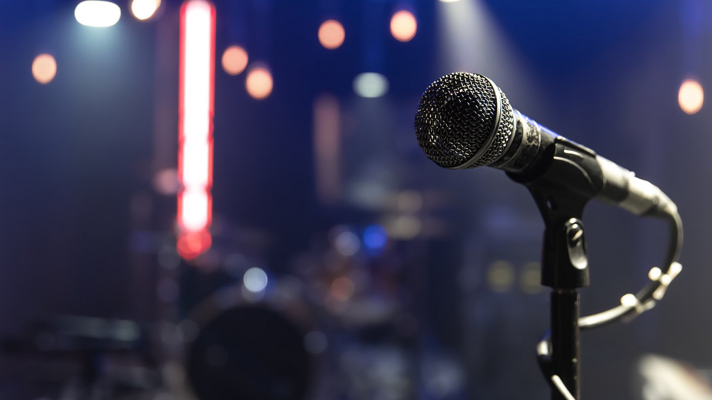 Das Bild zeigt ein Mikrofon vor einer Bühne mit atmosphärischer Beleuchtung.