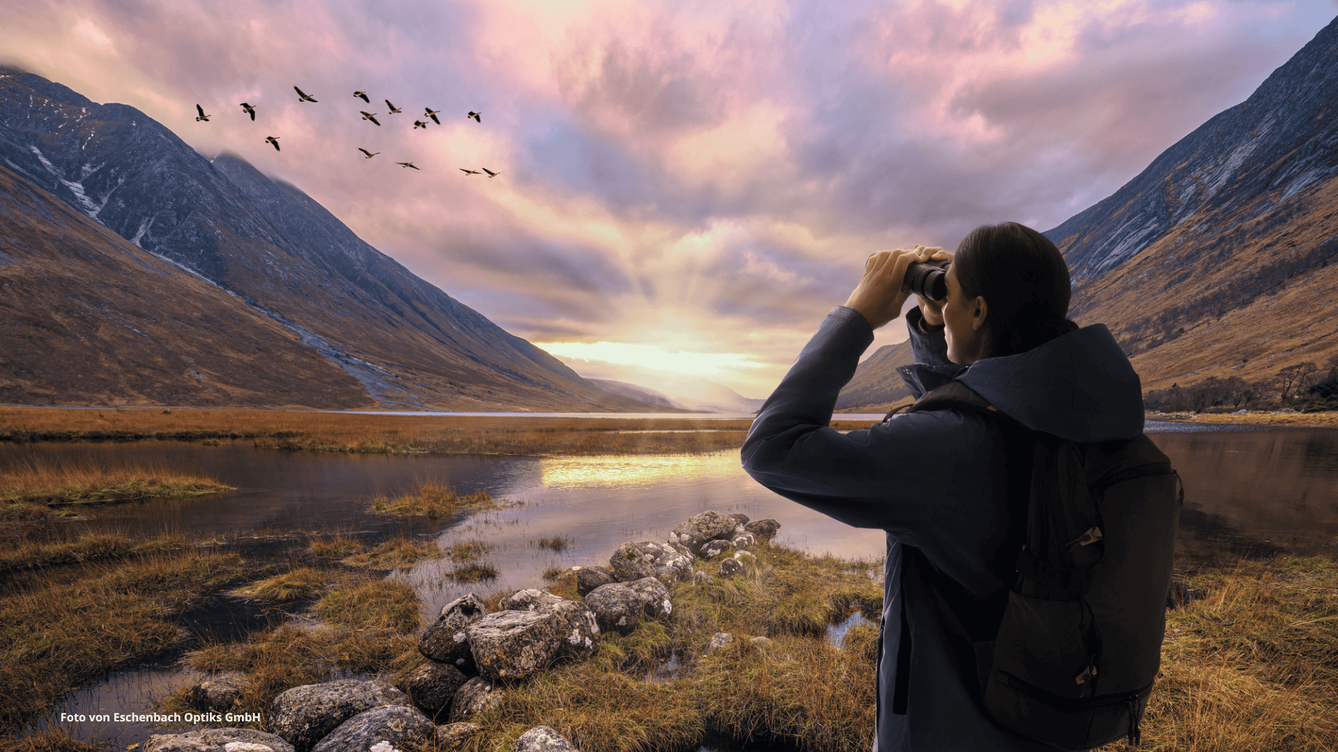 Das Foto zeigt eine Person, die bei Sonnenuntergang in einer Berglandschaft Vögel am Himmel mit einem Fernglas beobachtet.